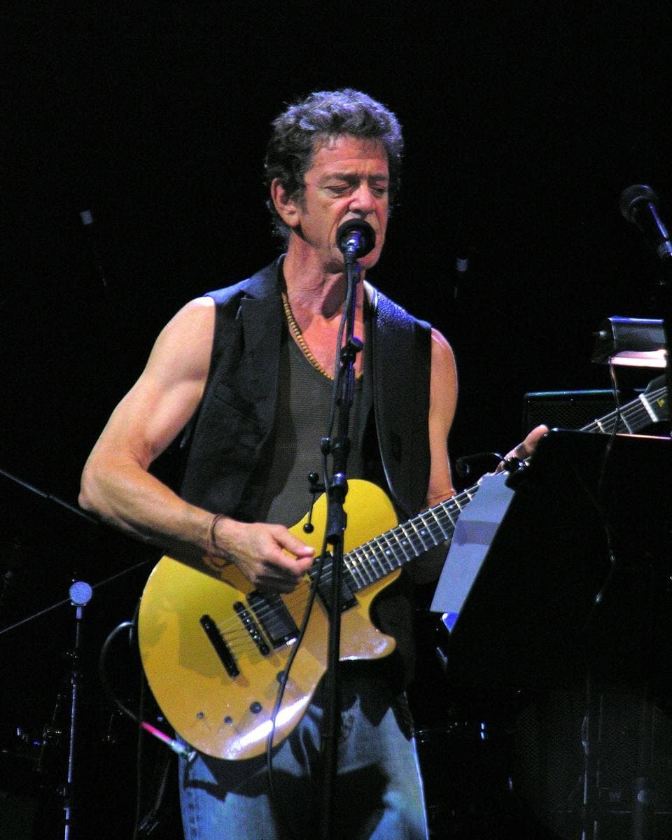 Lou Reed in a black waistcoat singing and playing a yellow guitar under stage light