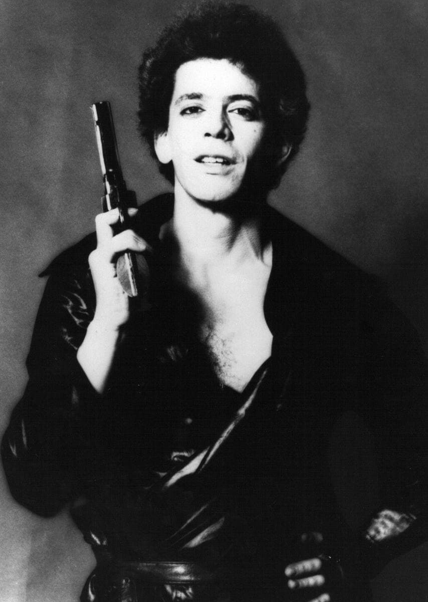Lou Reed in black and white, curly dark hair, open dark shirt, holding a prop pistol upright