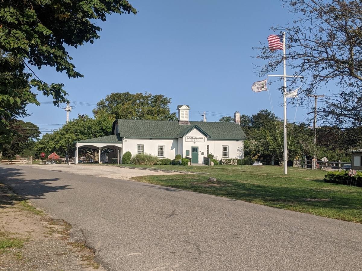 A small white clapboard hall with a green roof and a flagpole beside a quiet road