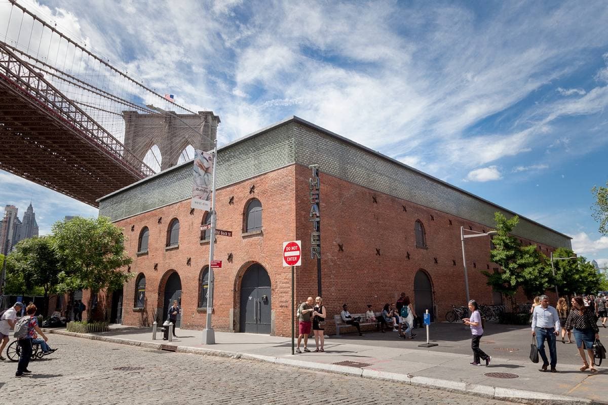 A long red-brick warehouse with arched doorways beneath the stone arch of the Brooklyn Bridge