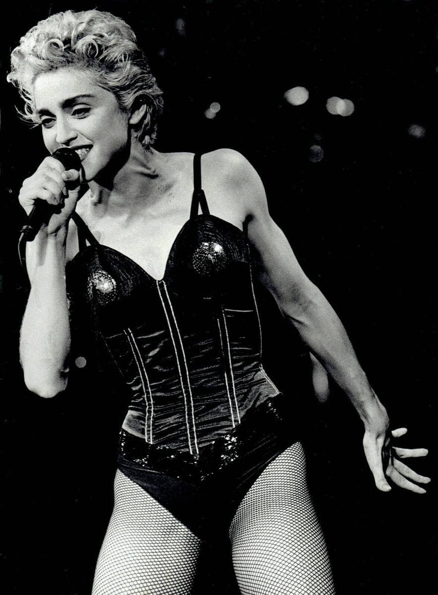 Madonna with short platinum curls singing into a handheld microphone in a corset and fishnets, black and white