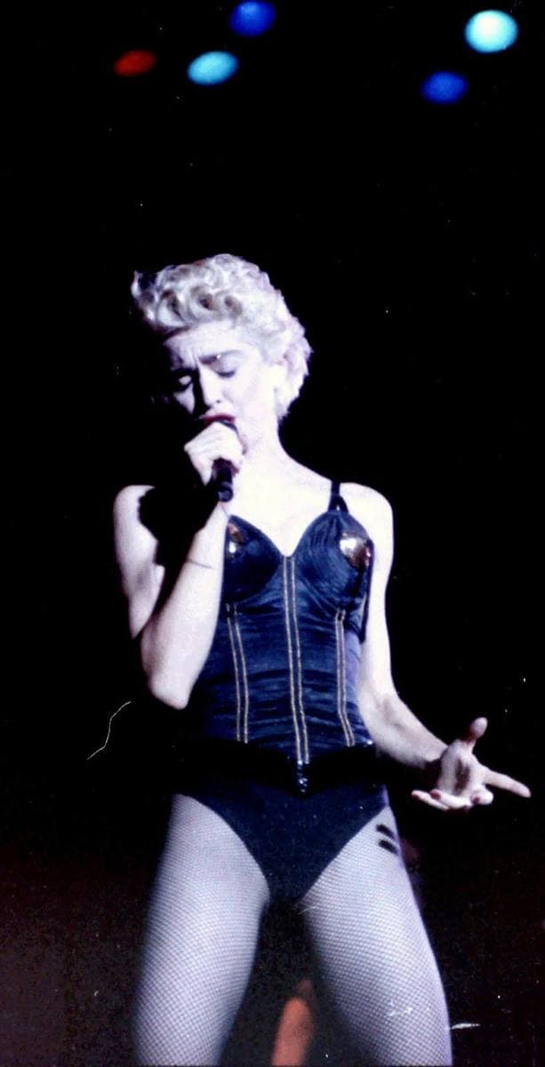 Madonna in a black corset under blue stage light, singing into a microphone