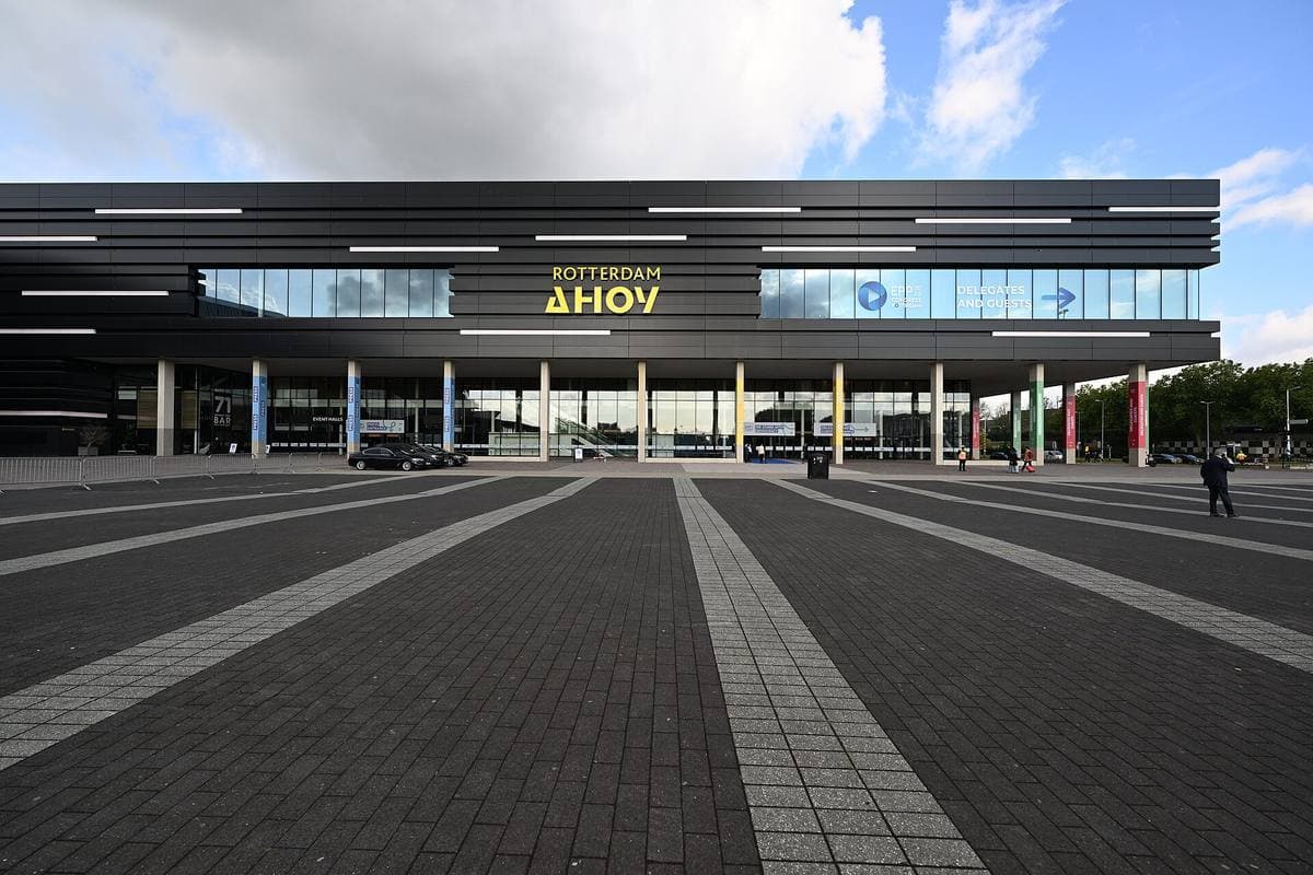 A long low black arena front with glass under a summer sky, a wide paved forecourt