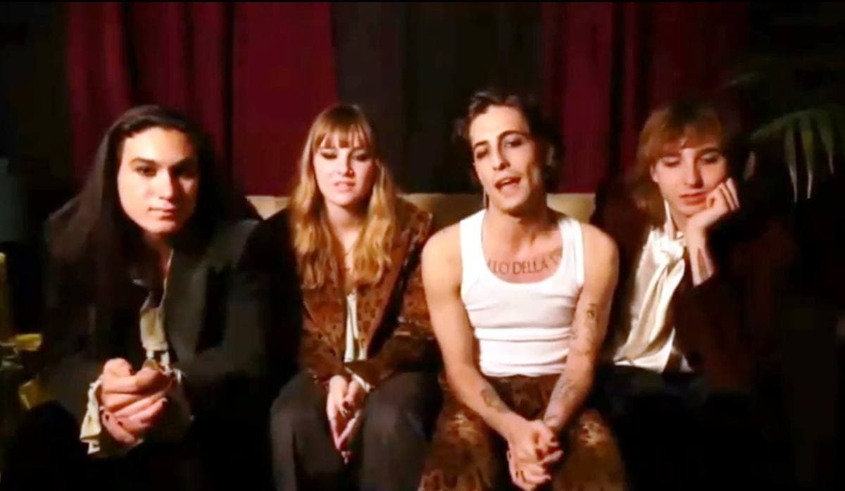 The four members of Måneskin sitting side by side on a sofa in front of a red curtain