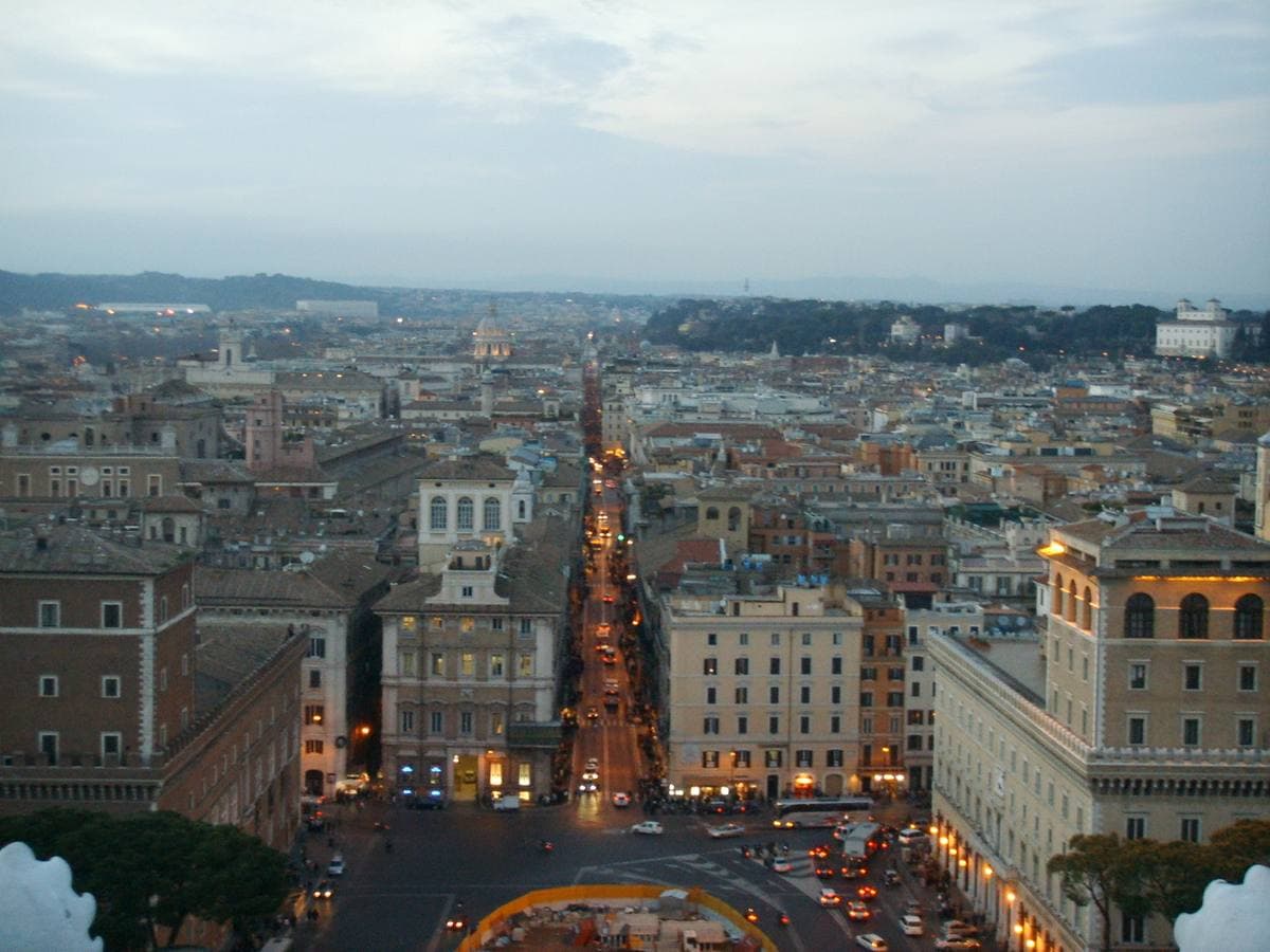 Looking down the long straight line of Via del Corso at dusk, lit shop fronts between tall palazzi