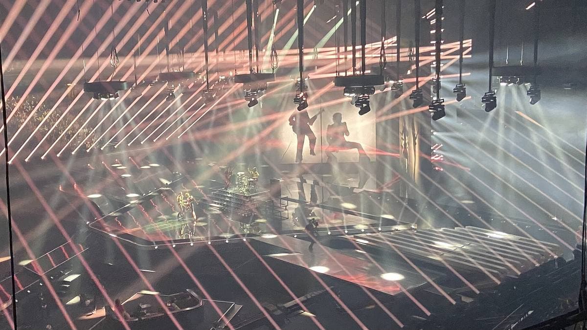 An arena stage criss-crossed by pink and white light beams, the band small at the centre