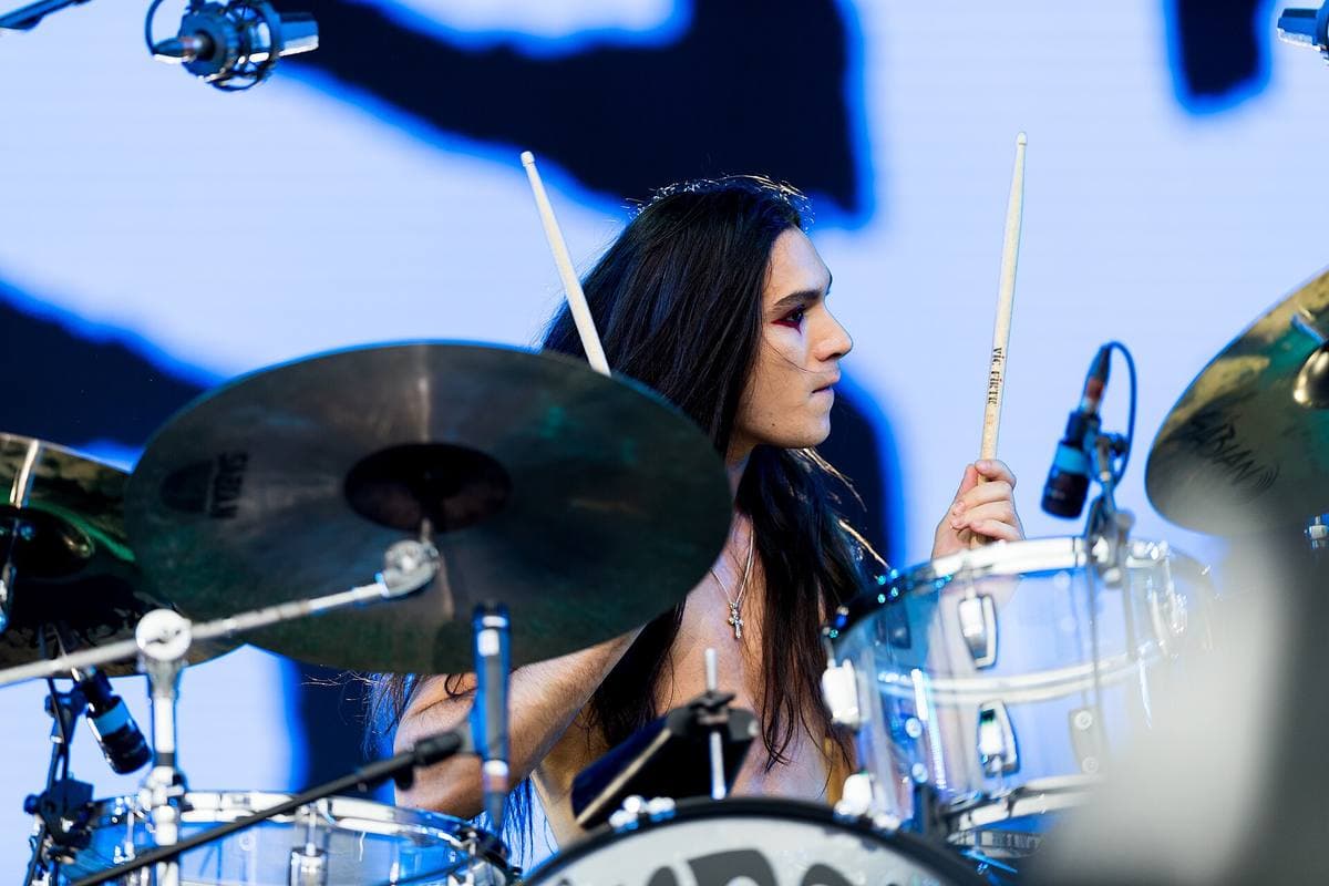 Ethan Torchio with long dark hair playing drums behind clear cymbals