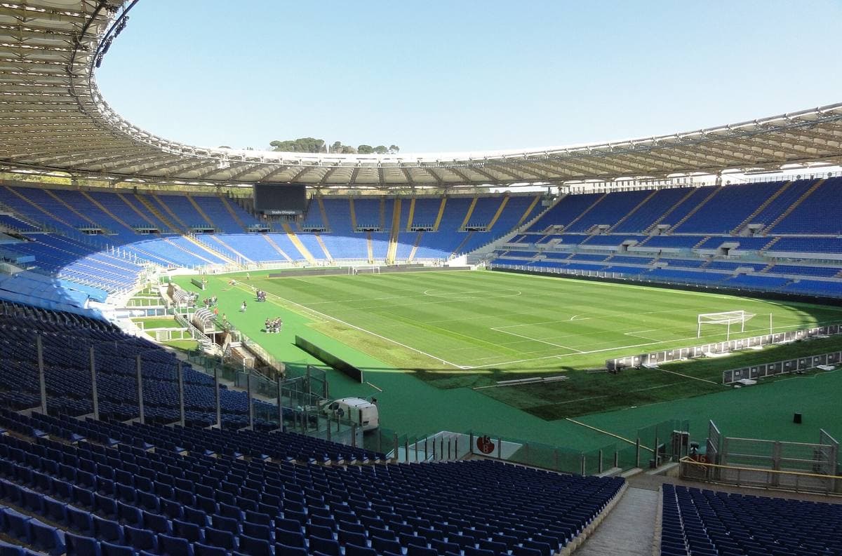 The bowl of a big stadium with blue seats and a green pitch under a clear sky
