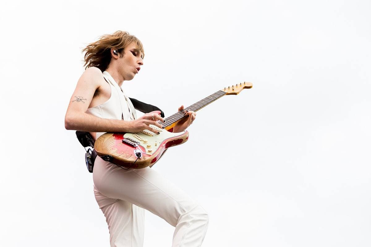 Thomas Raggi in white, leaping with a worn electric guitar against a pale sky