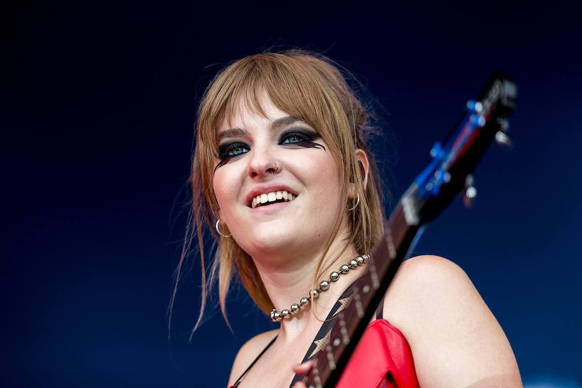 Victoria De Angelis smiling on stage, bass guitar neck across the frame, dark eye make-up