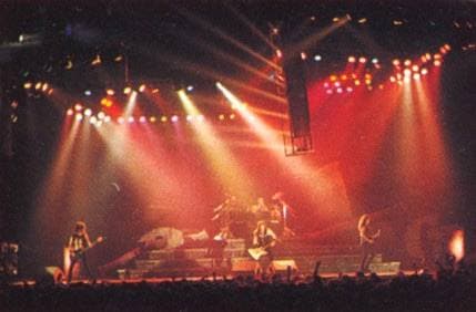A wide arena stage in red and white spotlights with the band small at the front