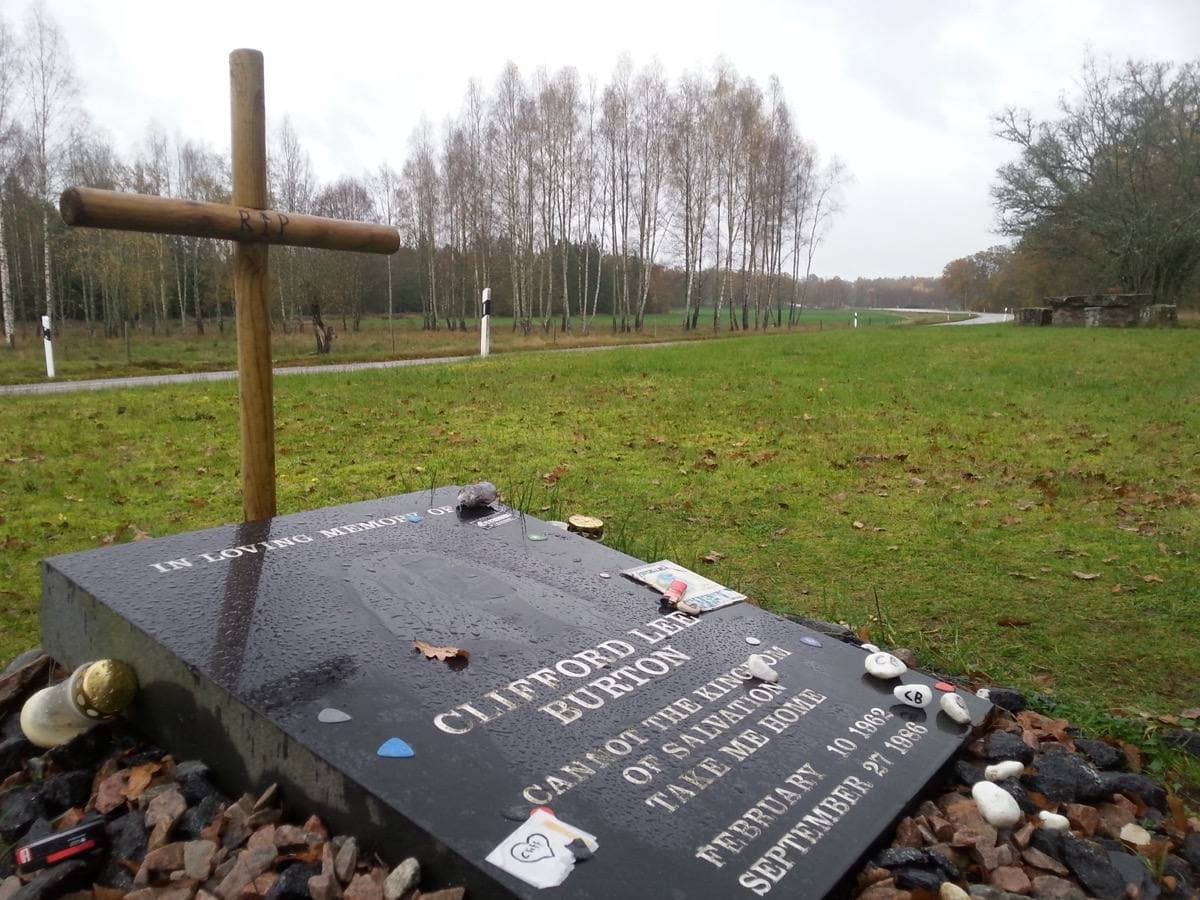 A rough grey memorial stone with an engraved plaque in a small clearing by a road