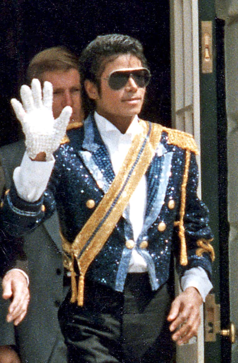 Michael Jackson in a blue sequinned jacket with a gold sash, raising a white sequinned glove