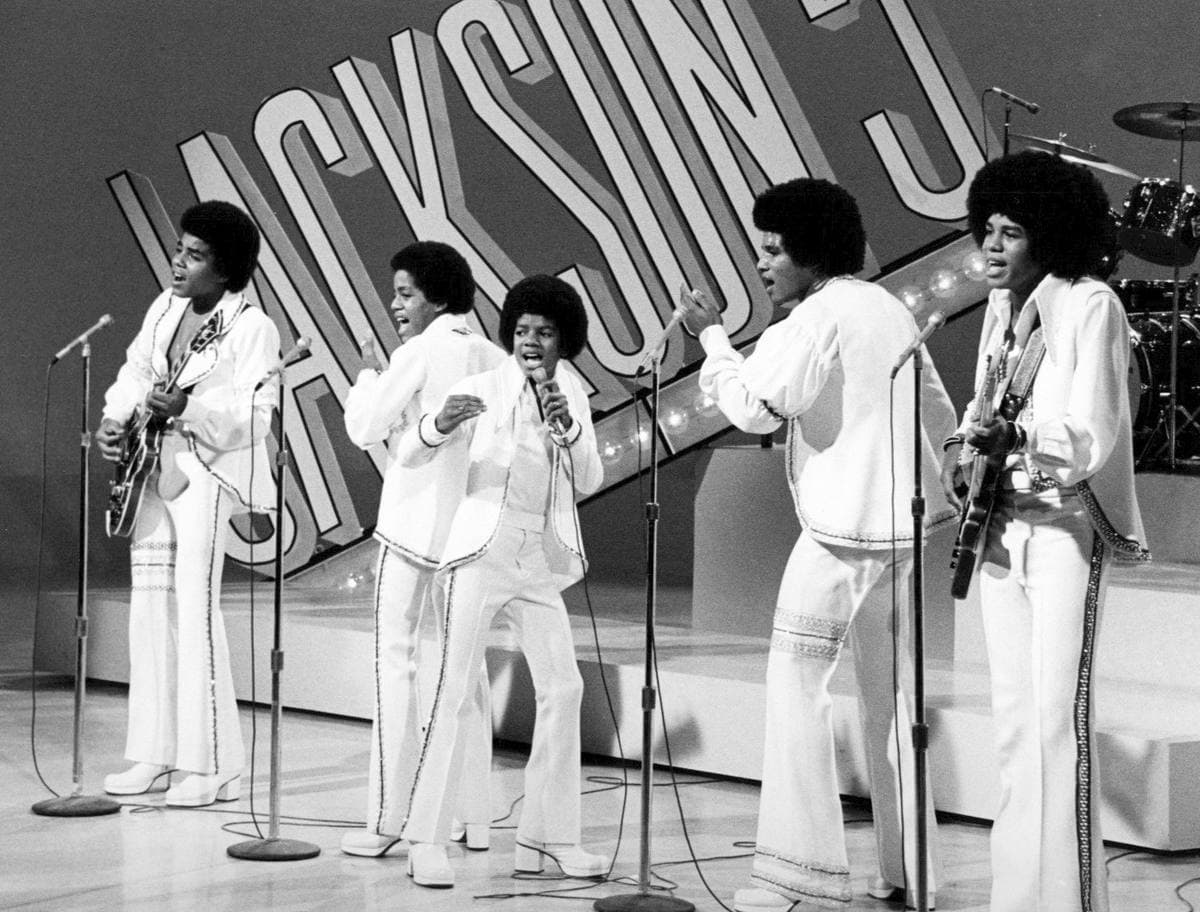 The five Jackson brothers in white suits singing at microphone stands in front of a giant Jackson 5 sign
