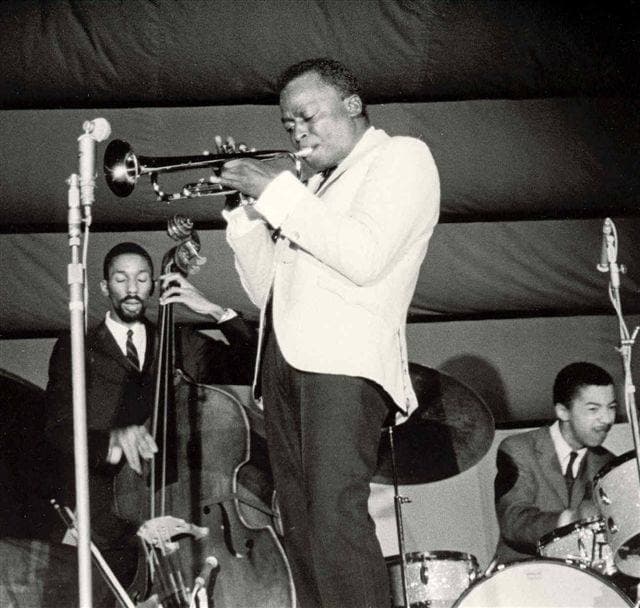 Miles Davis in a pale jacket plays trumpet into a microphone on stage, bass and drums behind him