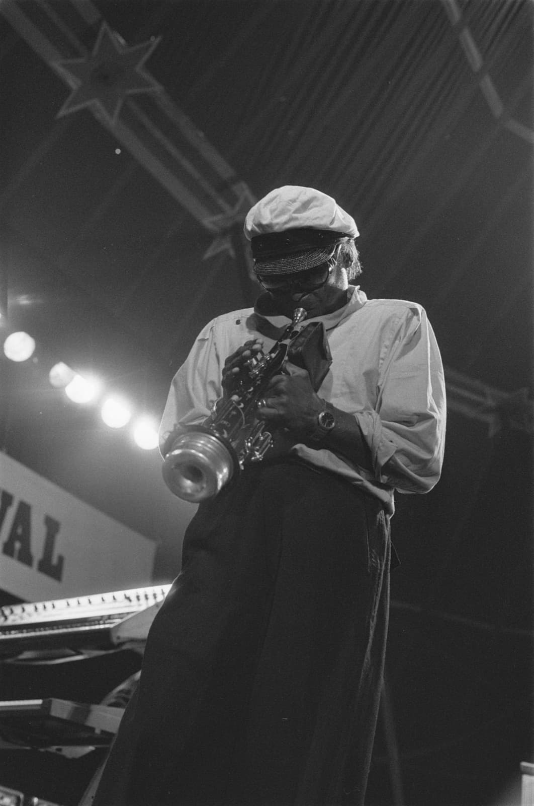 Miles Davis stands bent over his trumpet, the bell pointing down at the stage, lights behind him