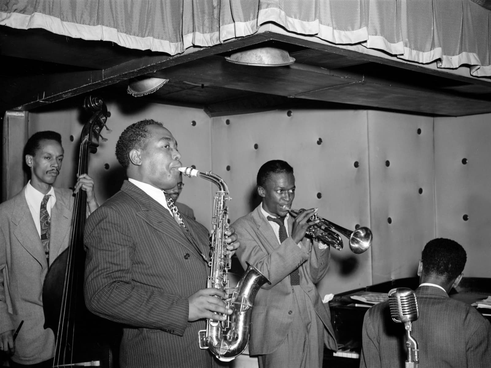 Miles Davis, aged 21, plays trumpet beside Charlie Parker on alto saxophone in a small padded club, with Tommy Potter on bass and Duke Jordan at the piano