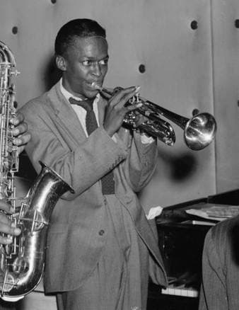 A young Miles Davis in a suit, playing trumpet in a club, a saxophone beside him