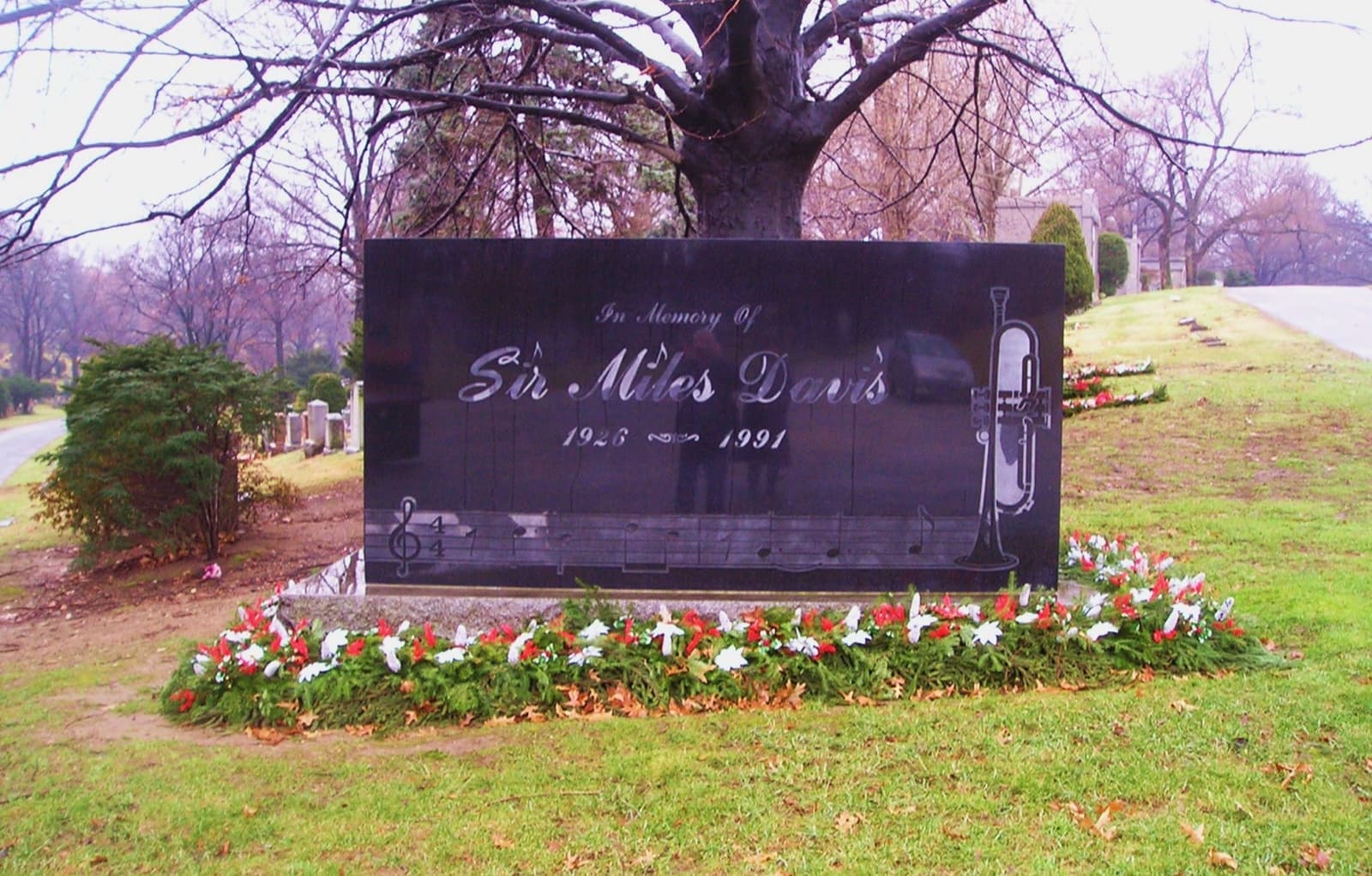 A polished black granite stone inscribed Sir Miles Davis, with flowers along its base and a tree behind