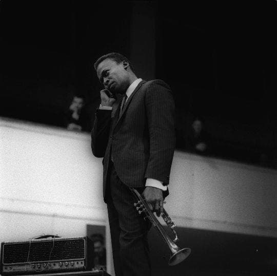 Miles Davis stands in shadow on a dark stage, trumpet held low at his side