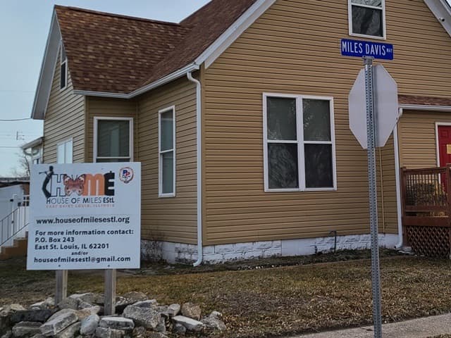 A small two-storey house with tan siding on a street corner, a House of Miles sign in the yard and a Miles Davis street sign