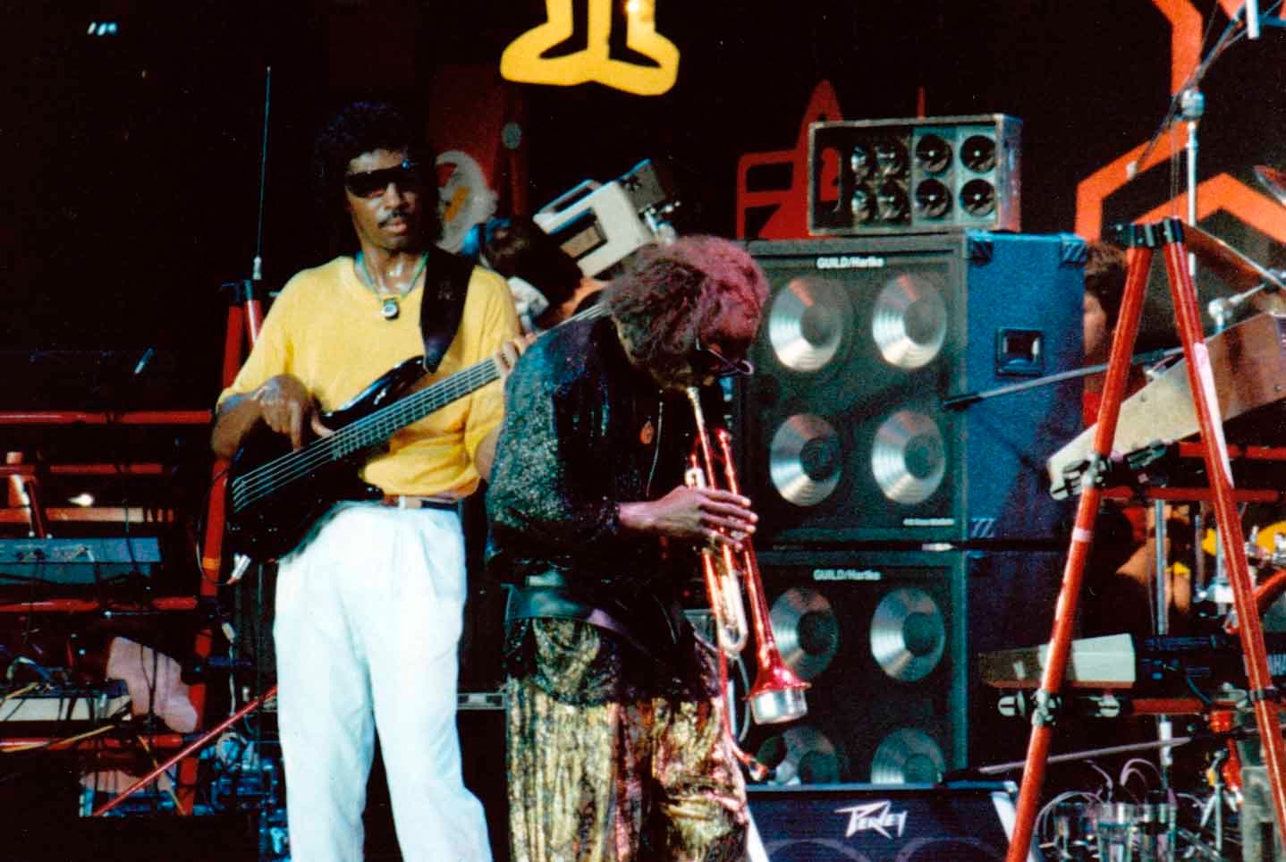 Miles Davis playing trumpet on a crowded stage beside a bassist in a yellow shirt