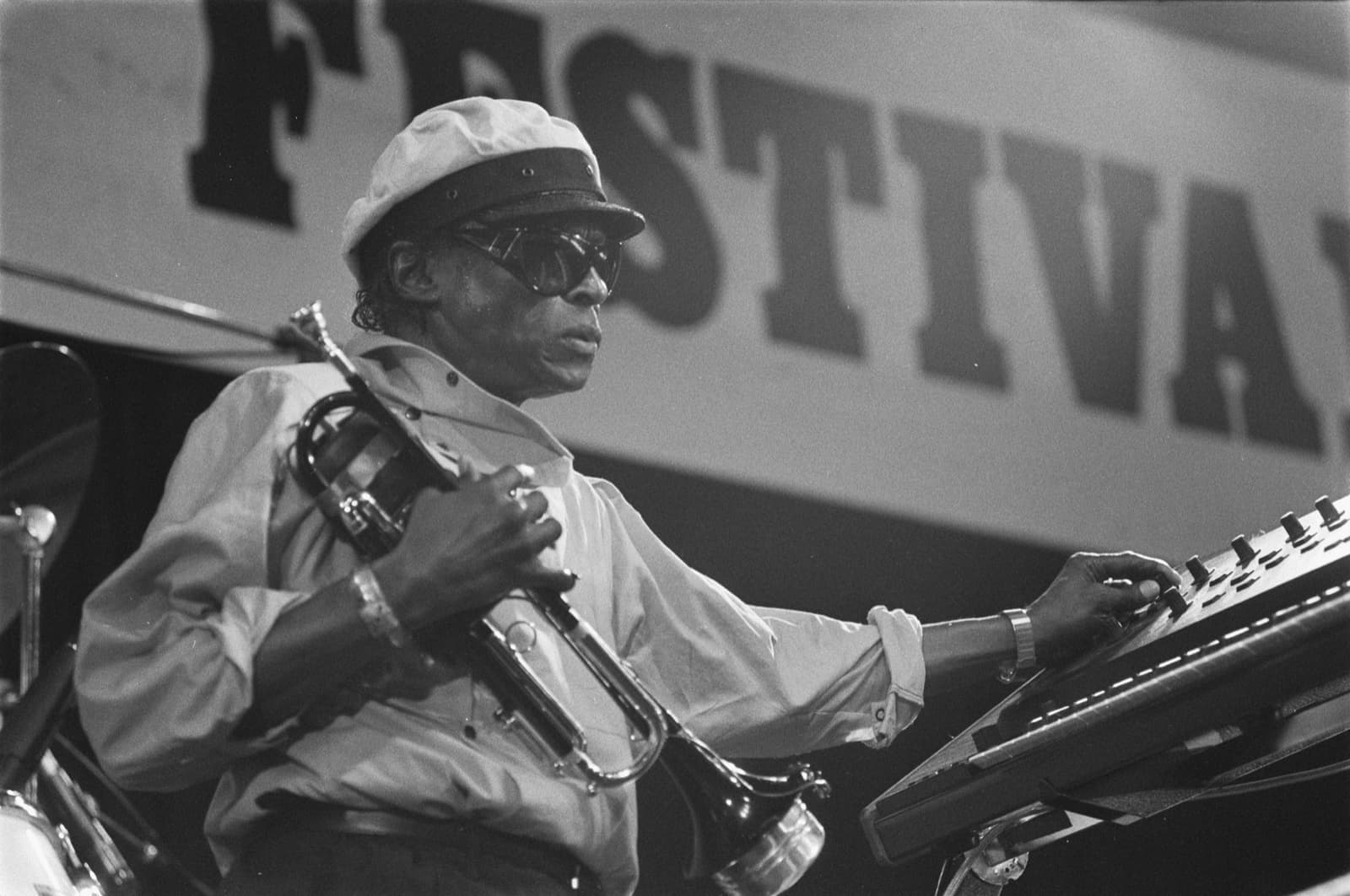 Miles Davis in a white cap and dark glasses holds his trumpet and plays a keyboard on stage