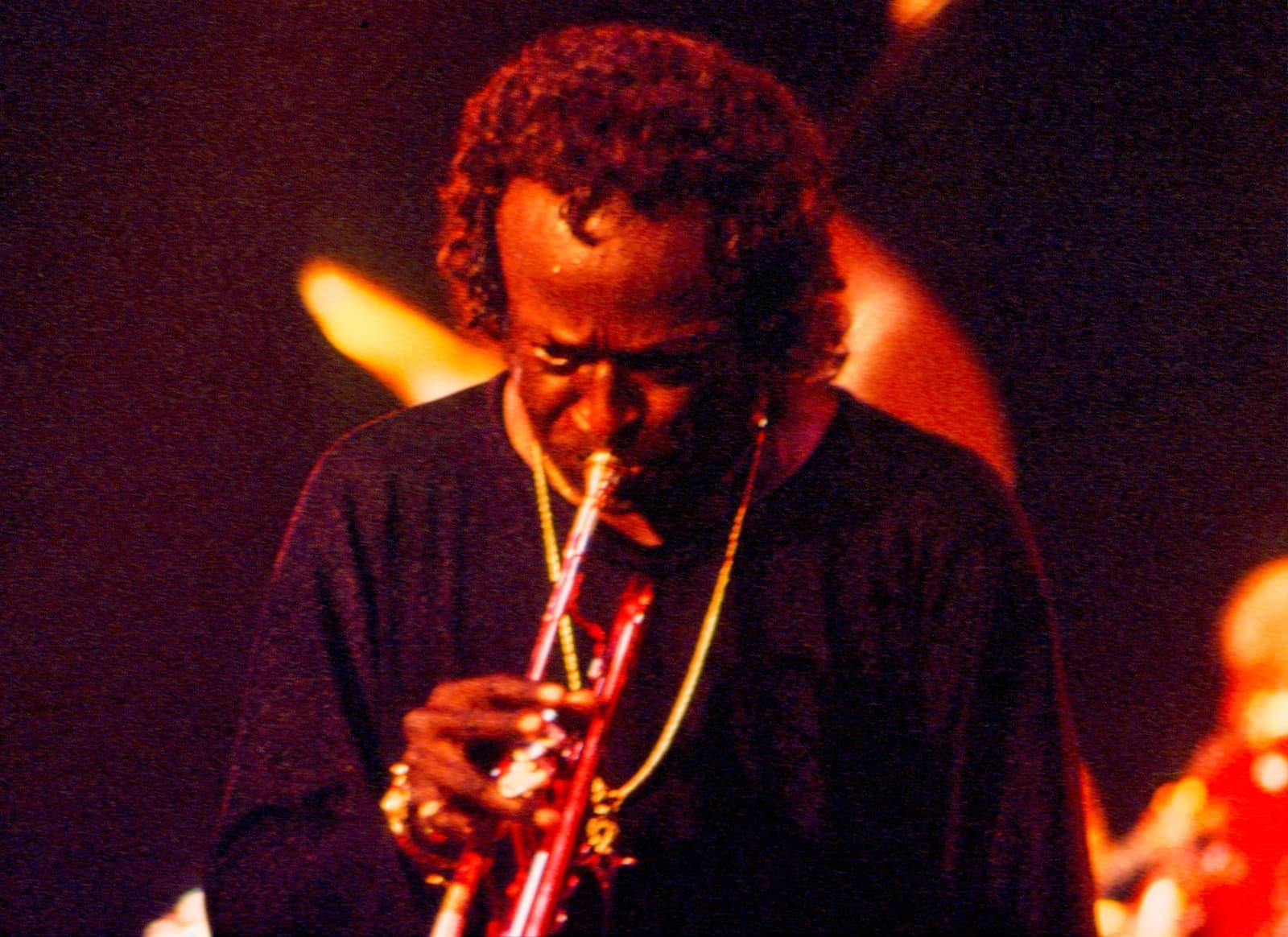 Miles Davis in close-up, eyes down, playing a red trumpet under warm stage light
