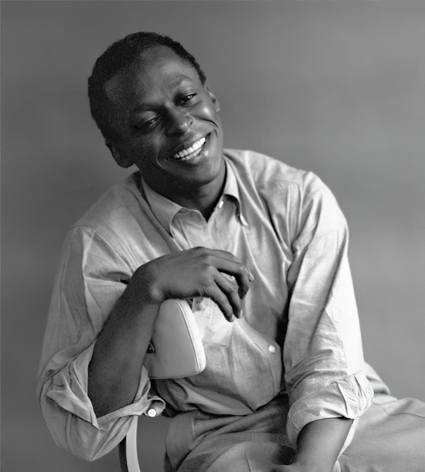 Miles Davis seated in an open-necked shirt, laughing, his hands folded over his knee