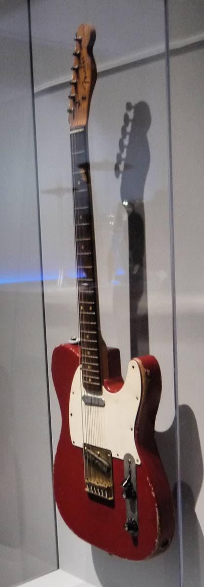 A worn red Fender Telecaster with a cream scratchplate standing upright in a glass case