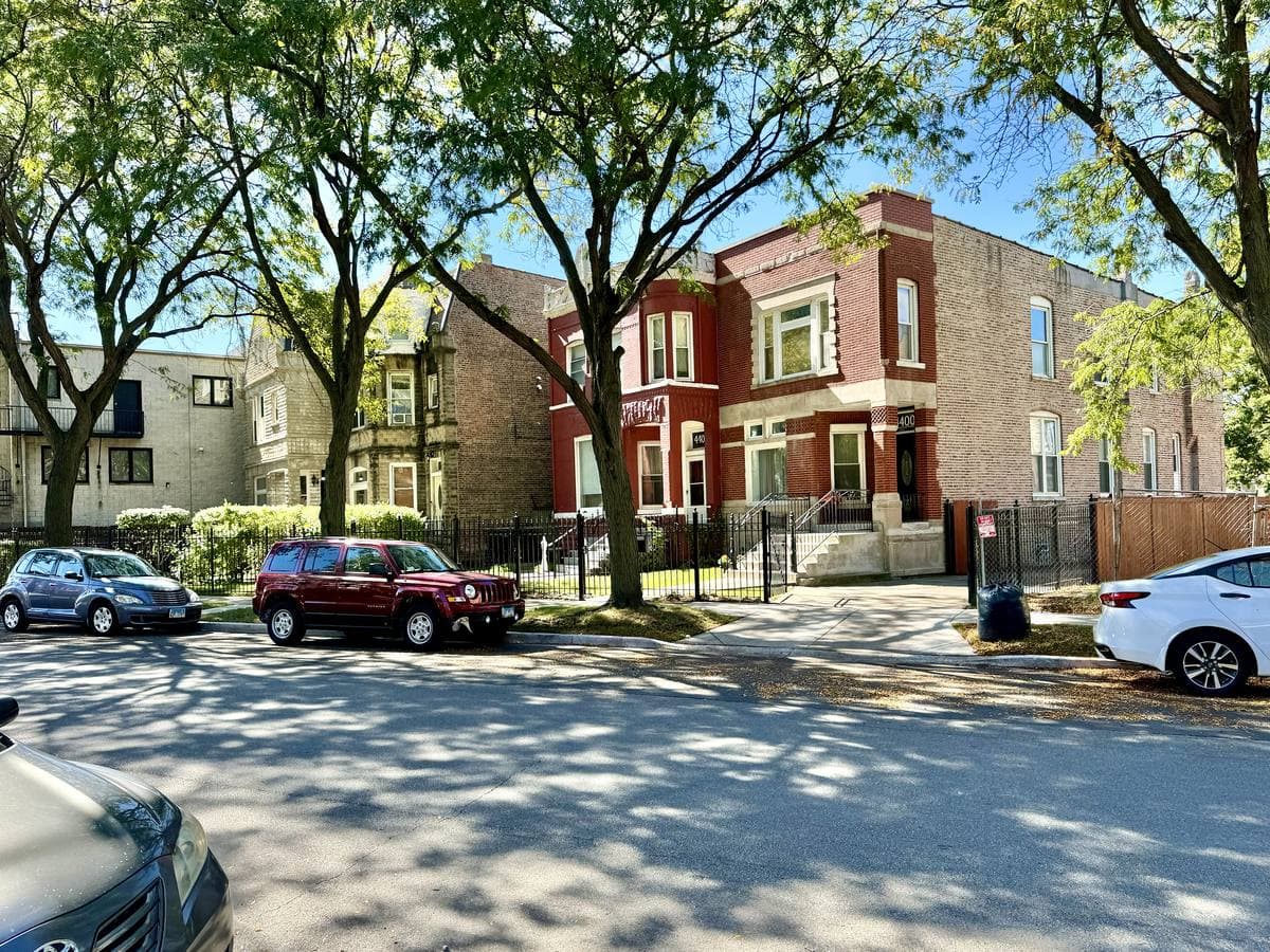 A tree-lined residential street of brick two-flats with cars parked along the kerb