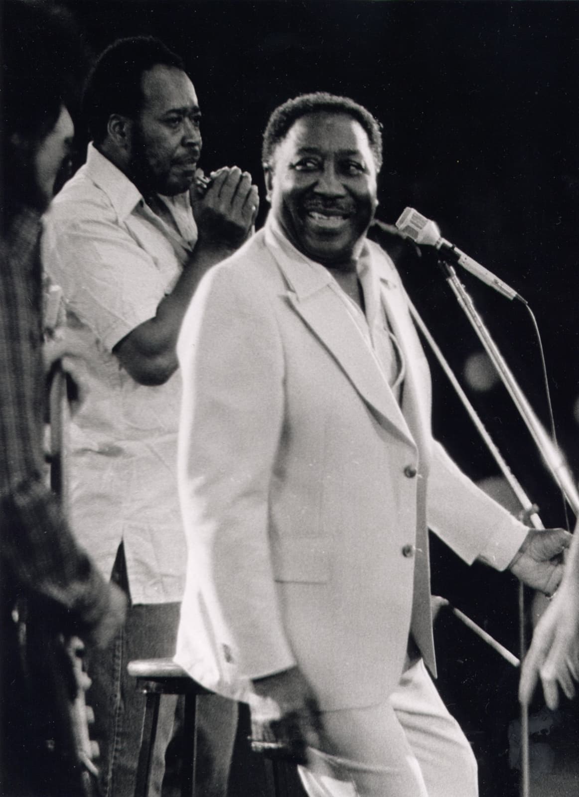 Muddy Waters in a white suit laughing on stage beside a harmonica player