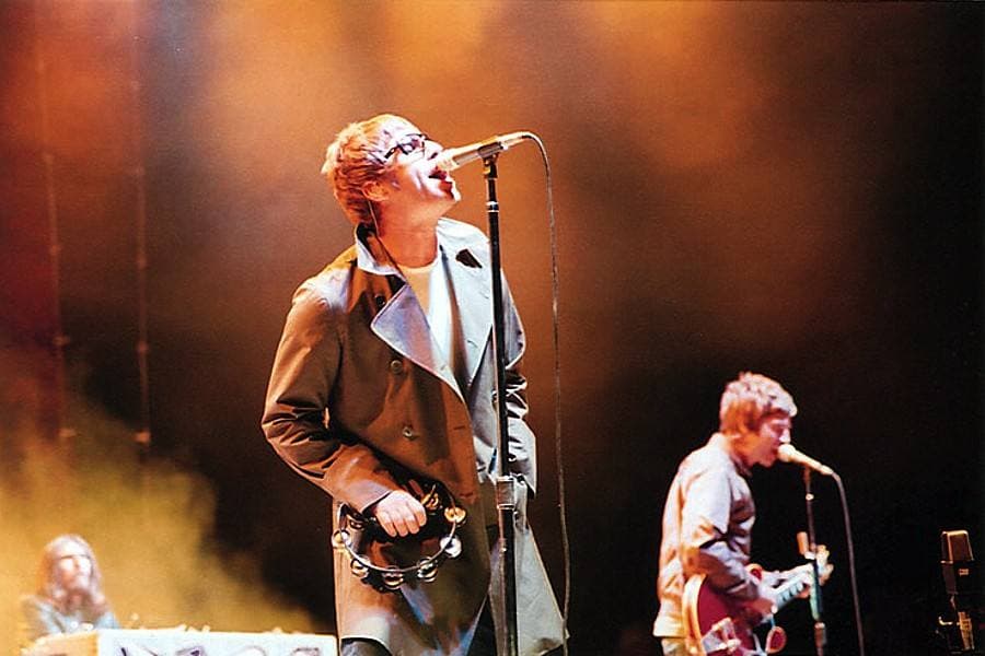 Liam Gallagher in a parka at the microphone with Noel Gallagher on guitar behind him, under orange stage light