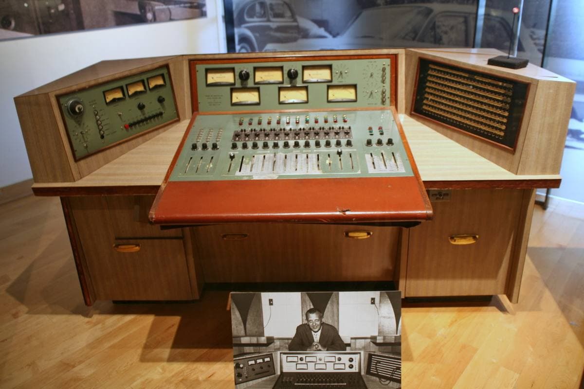 A wooden recording console with rows of knobs and meters on a museum floor