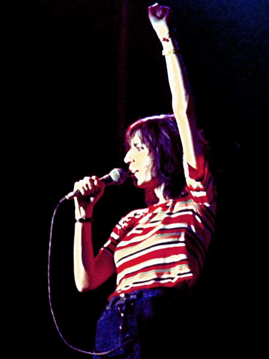 Patti Smith in a red and white striped top, one arm raised high, singing into a microphone against black