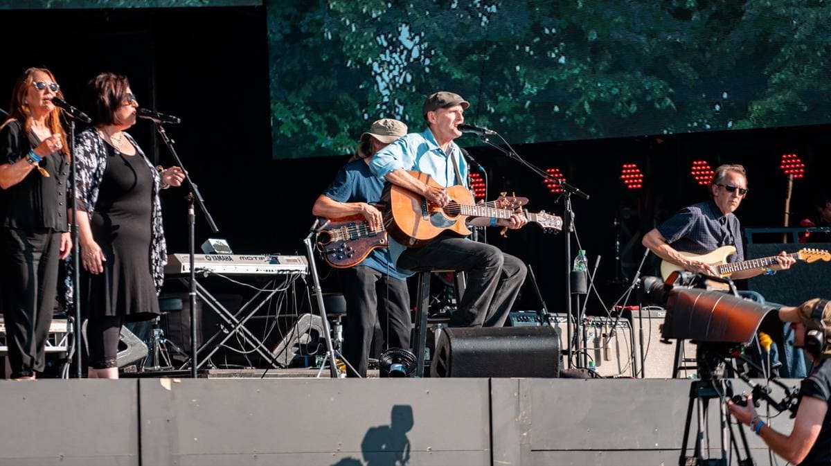 Paul Simon in a hat playing acoustic guitar on a big outdoor stage with his band