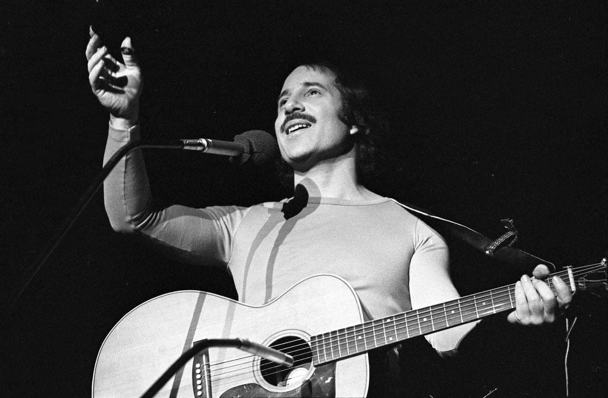 Paul Simon singing at a microphone with an acoustic guitar, one arm raised