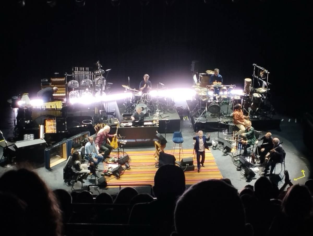 A concert stage lit white from behind, the band seated in a half circle