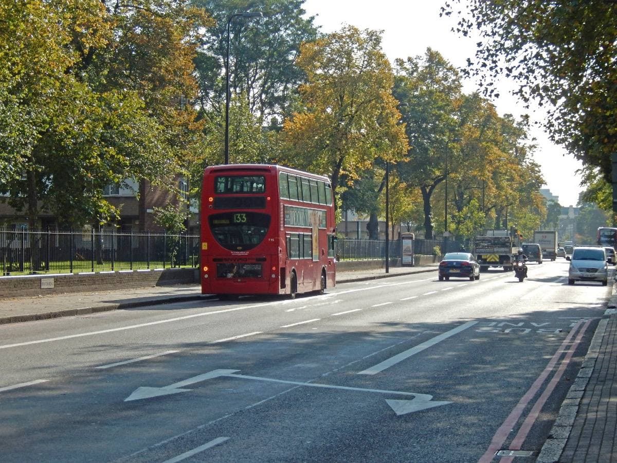 A red double-decker bus on a wide tree-lined Brixton Road