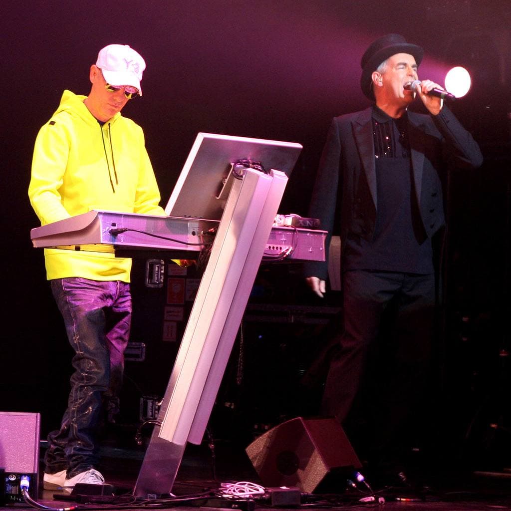 Chris Lowe in a yellow top and white cap at a keyboard stand, Neil Tennant in a dark suit singing, under magenta stage light