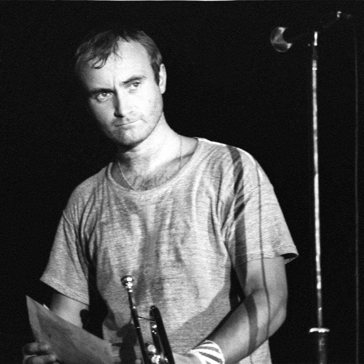 Phil Collins in black and white, holding a sheet of paper beside a microphone stand, looking at the camera