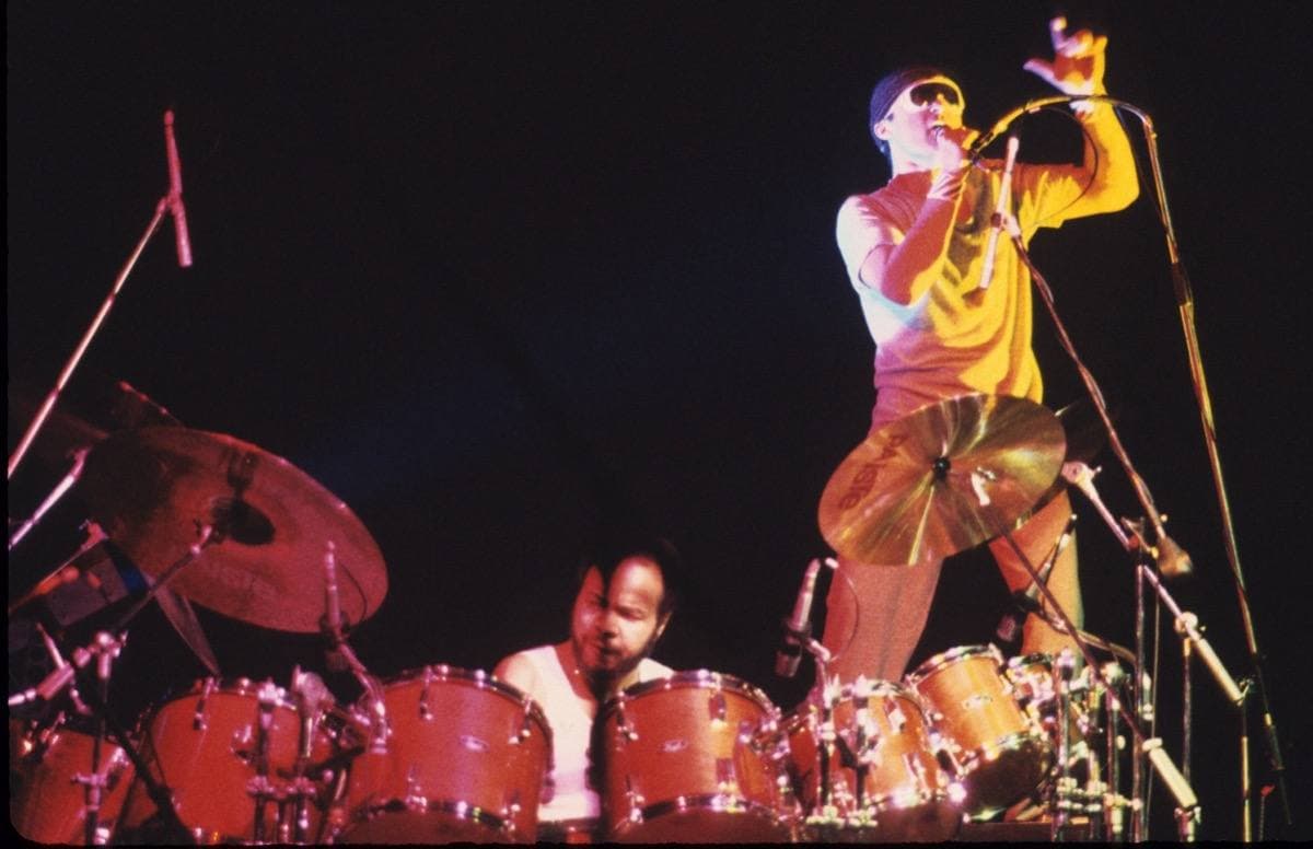 A drum kit in red light with Chester Thompson behind it and Phil Collins standing at a microphone