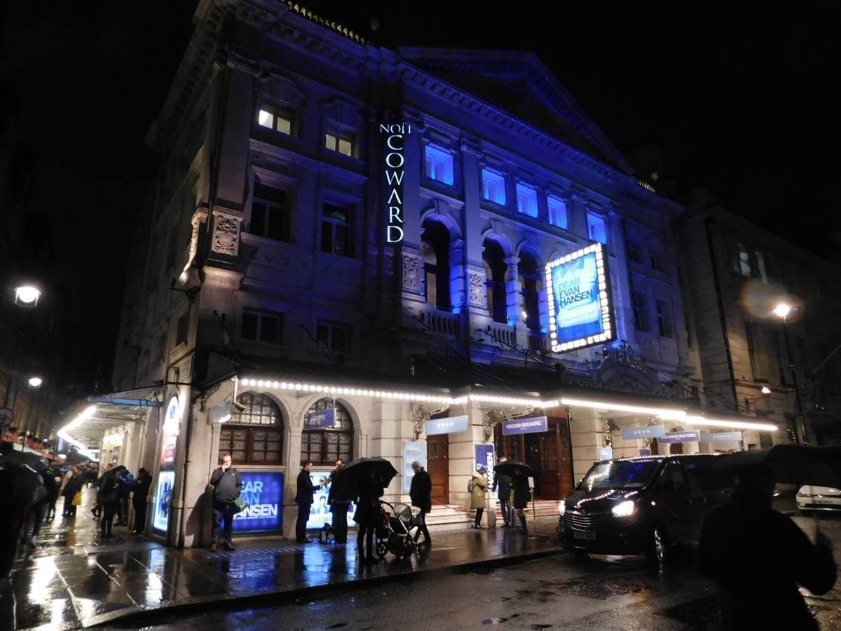 A theatre front on a wet night, lit blue, with people under the canopy