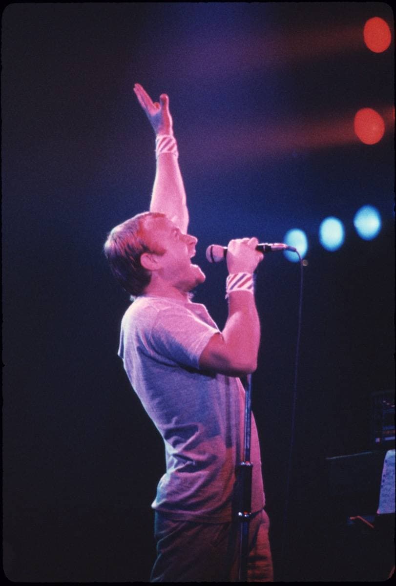 Phil Collins on stage in a grey T-shirt, one arm stretched up, blue and red lights behind