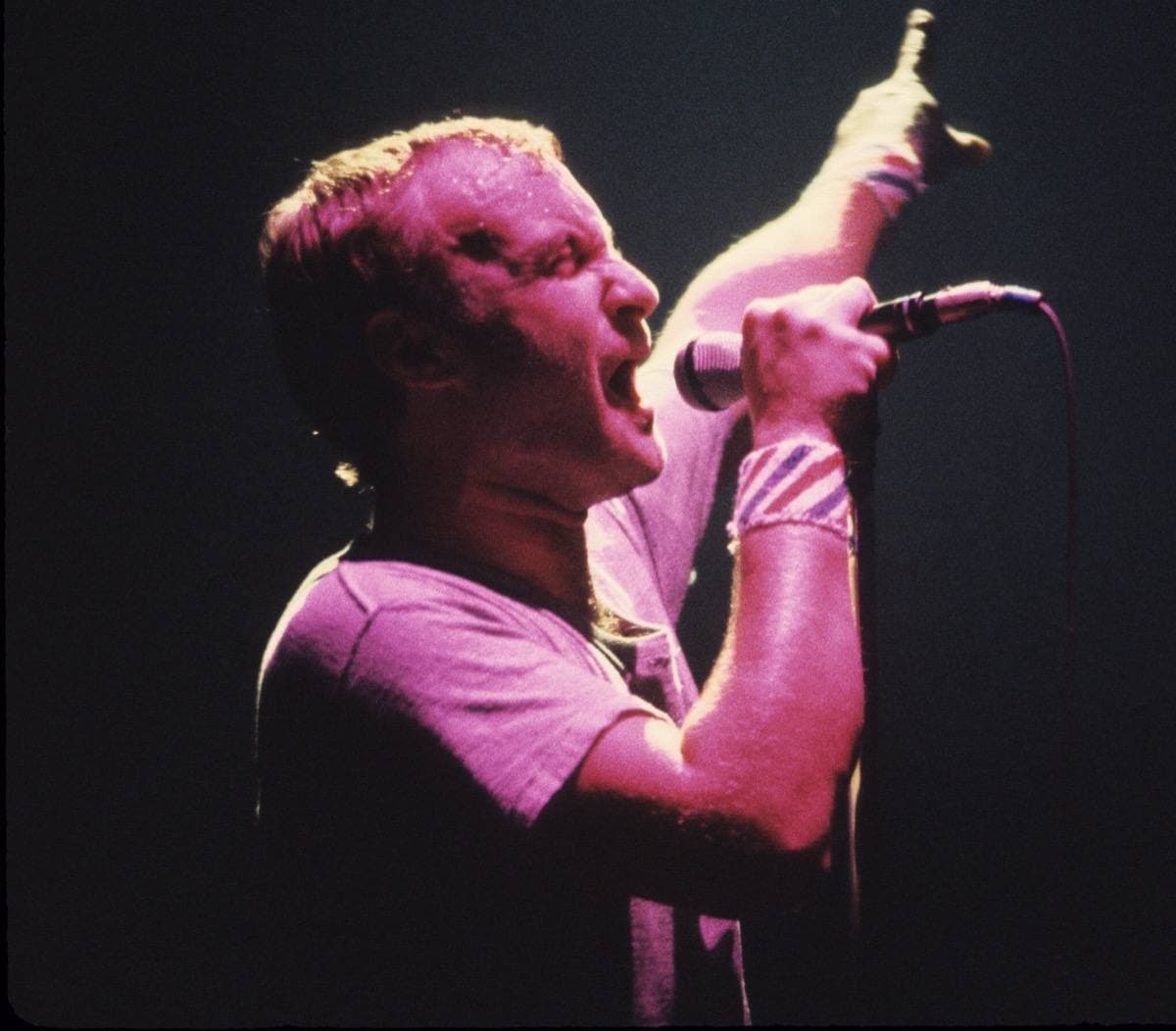 Phil Collins singing into a microphone under pink stage light, one arm raised
