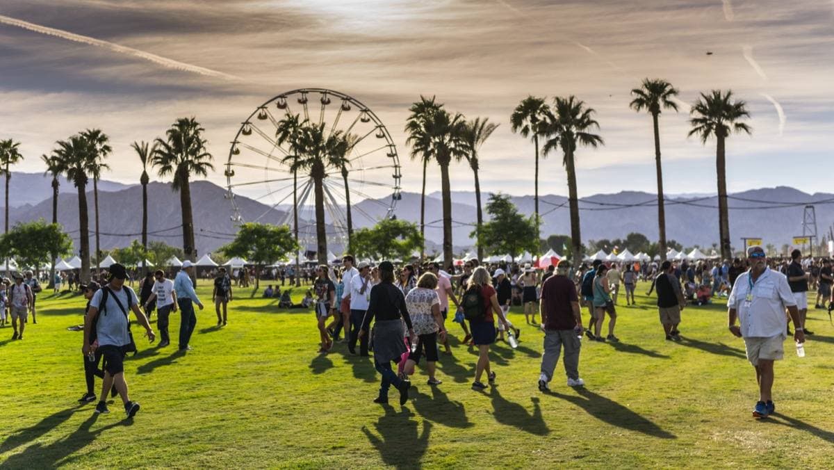 A crowd crossing a green field under palm trees, a Ferris wheel and mountains behind