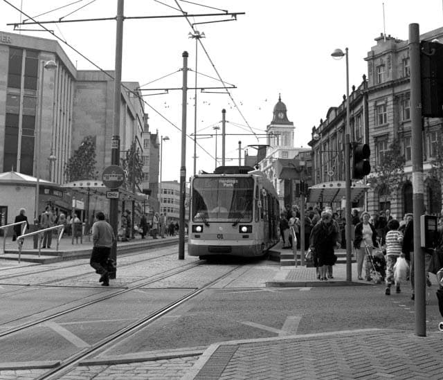 Black-and-white photograph of a Supertram stopped at Castle Square, Sheffield, in 1995, shoppers crossing the tracks under the overhead wires