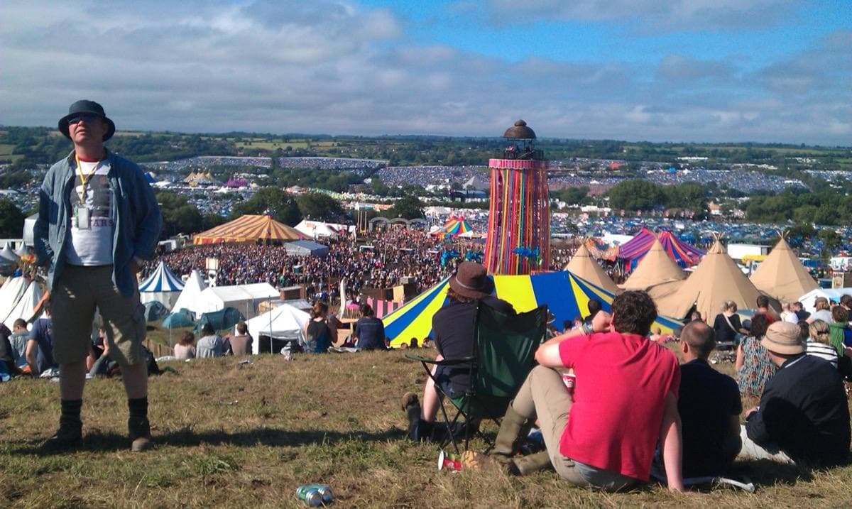 Glastonbury from the slope above the Park Stage in June 2011: a man in a hat stands over people sitting on the grass, tents and the crowd spread out below