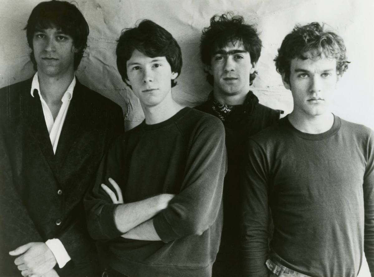 Four young men in dark shirts standing close together against a pale wall, black and white