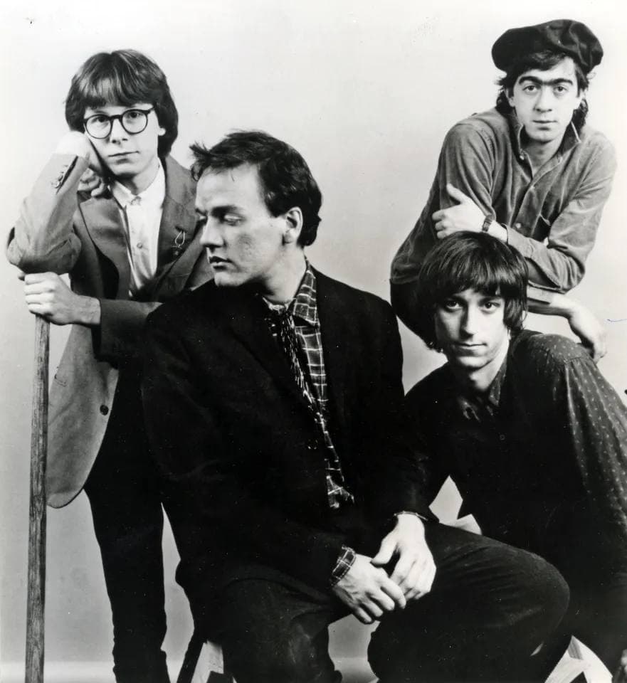 Four young men posed on and around a stool, one in round glasses, one in a beret, black and white